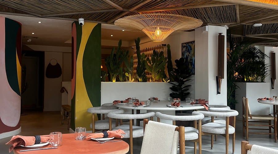 Interior of a restaurant with modern decor, wooden ceiling, green plants, and tables set with napkins and glassware.