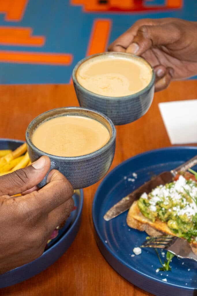 Two hands holding cups of coffee above plates with food on a wooden table