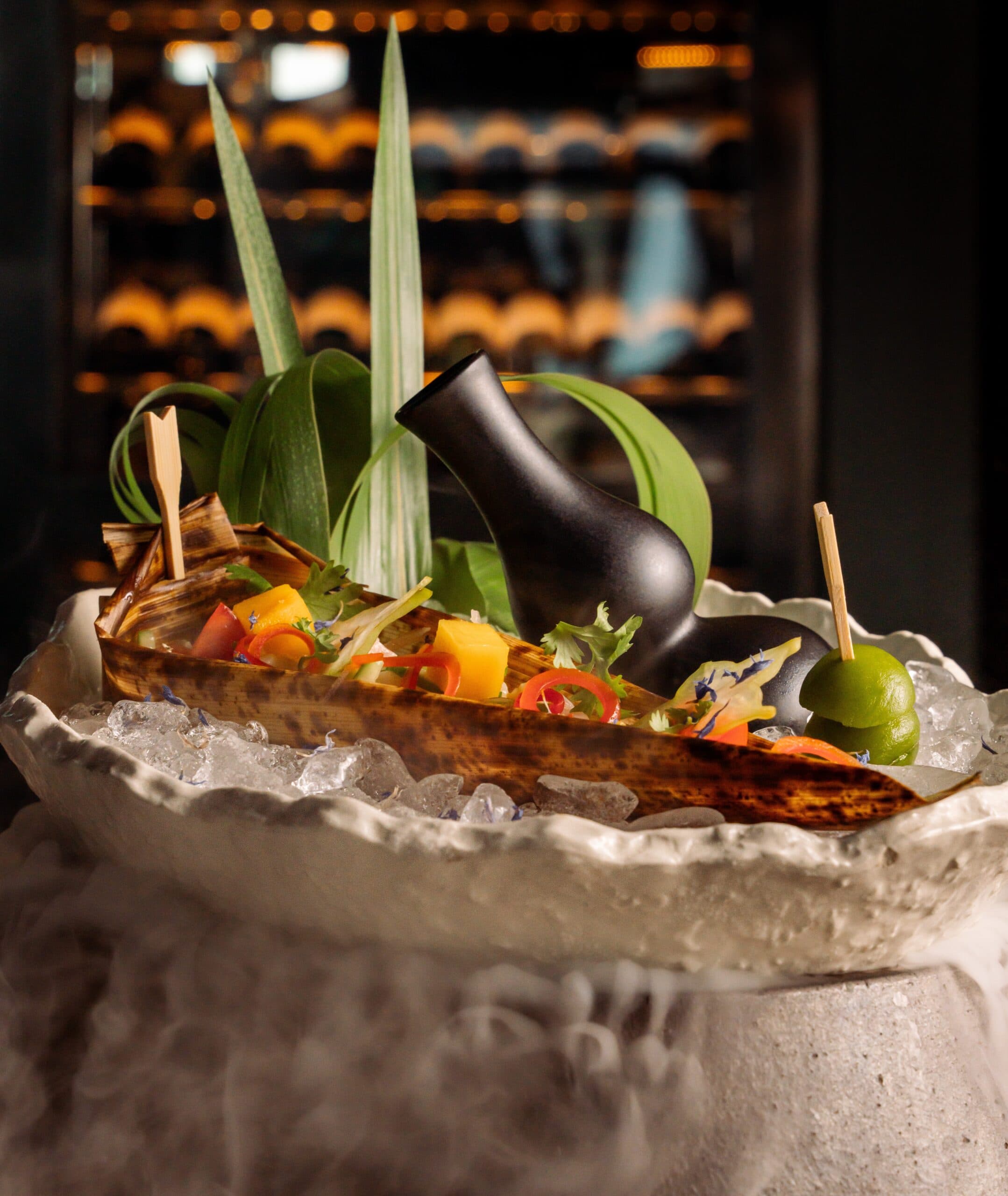 A beautifully presented dish with colorful ingredients on a bed of ice, featuring a rustic wooden container and a small black pitcher, surrounded by decorative leaves.